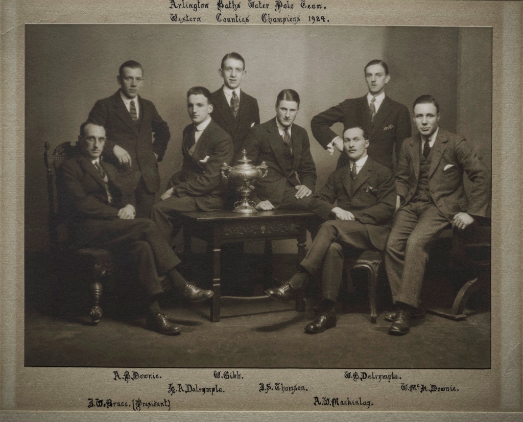 Black and white photo of eight young men in suits sitting and standaing around a table on which is standing a large silver trophy.